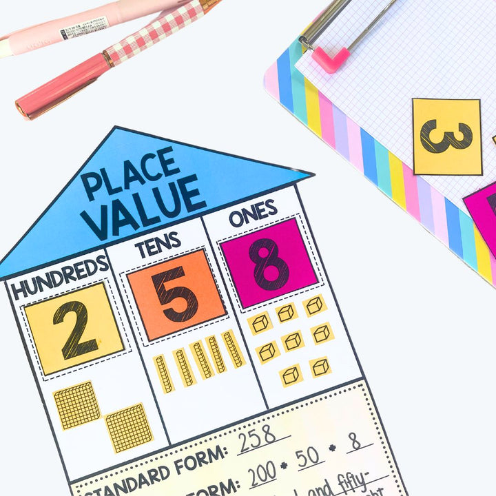 A colorful place value house activity for the number 258, placed on a desk with a rainbow clipboard, pens, and number cutouts.