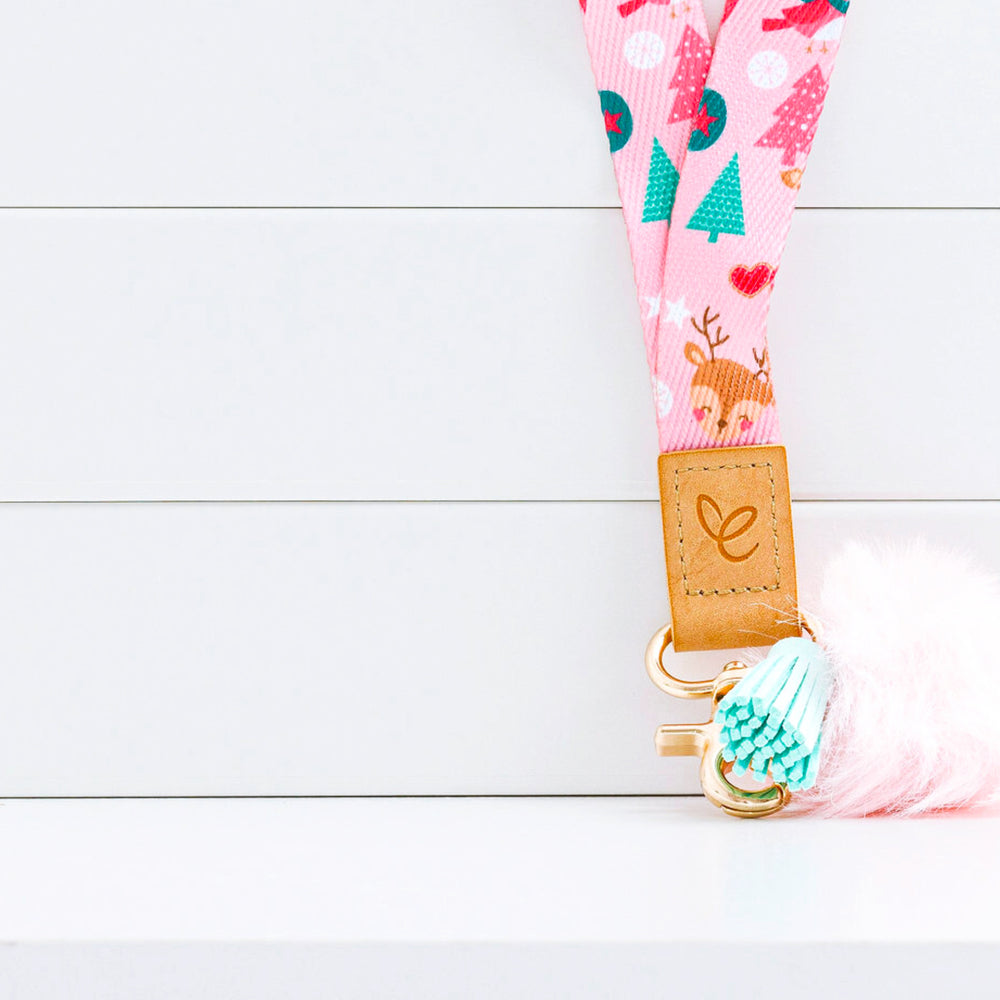 Close-up of a pastel pink Christmas lanyard featuring cute Santas, reindeers, snowmen, Christmas trees, and hearts. Includes a light pink pom-pom, aqua tassel, gold swivel clasp, and a light brown faux leather patch displaying the 'Mrs Edgar' logo.