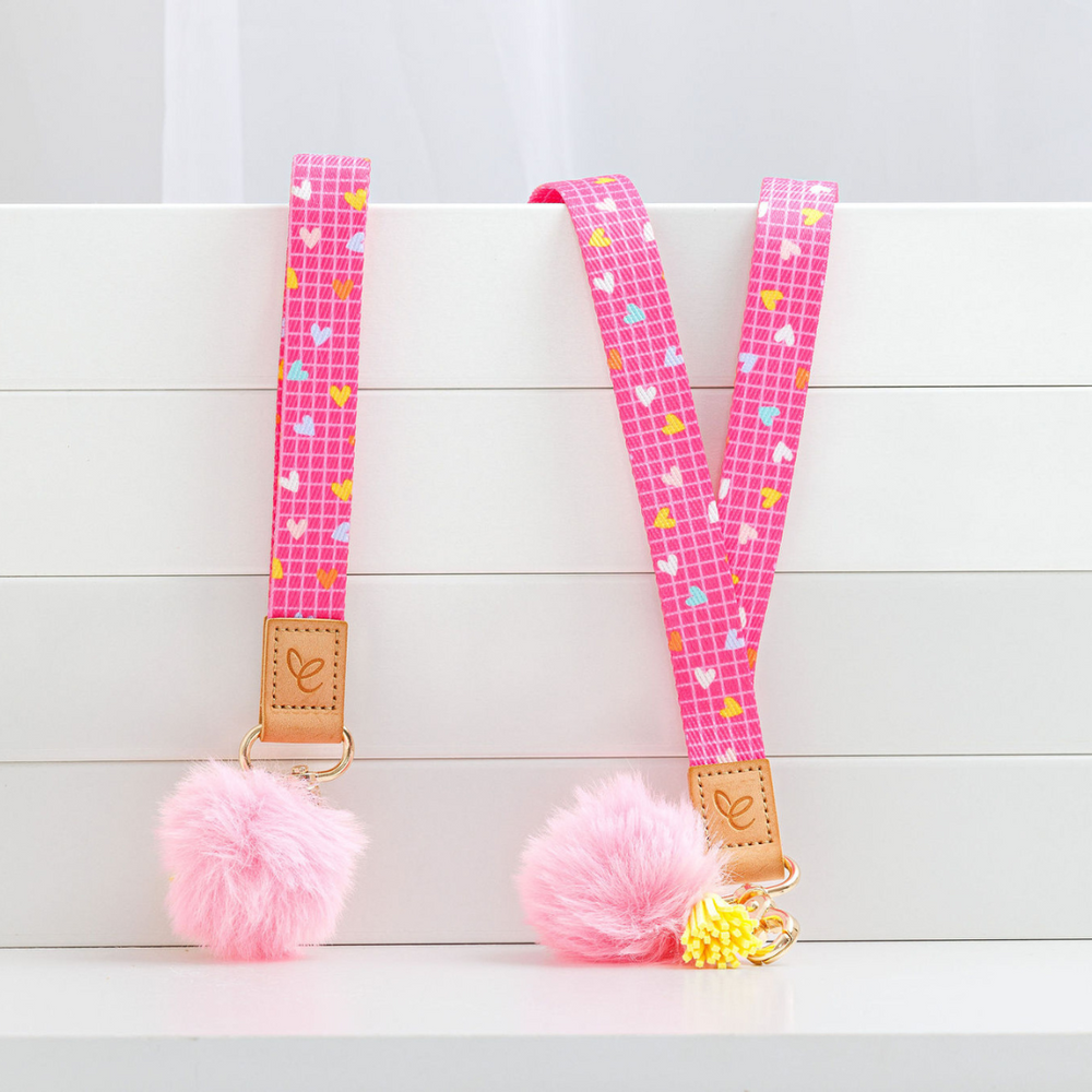 Bright pink lanyard and matching wristlet with white grid and heart pattern, featuring a light pink pom-pom, yellow tassel, and gold swivel clasp. A light brown faux leather patch displays the 'Mrs Edgar' logo.