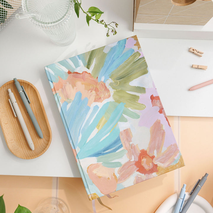 025 Classic Weekly Teacher Planner named Soirée, lying on a desk, showcasing earthy flora artwork created with paint strokes.