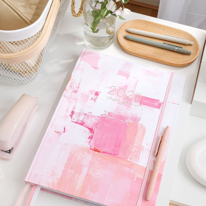 2025 Classic Weekly Teacher Planner in Love Vibes design, featuring pink and orange brush strokes, laying on a desk.