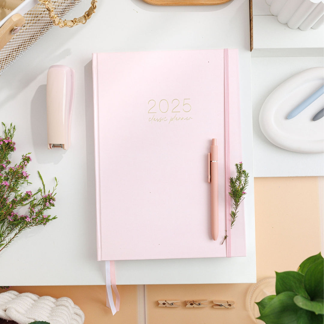 2025 Classic Weekly Teacher Planner in blush pink with gold text, coil-free, lying on a desk with a pen resting on top.