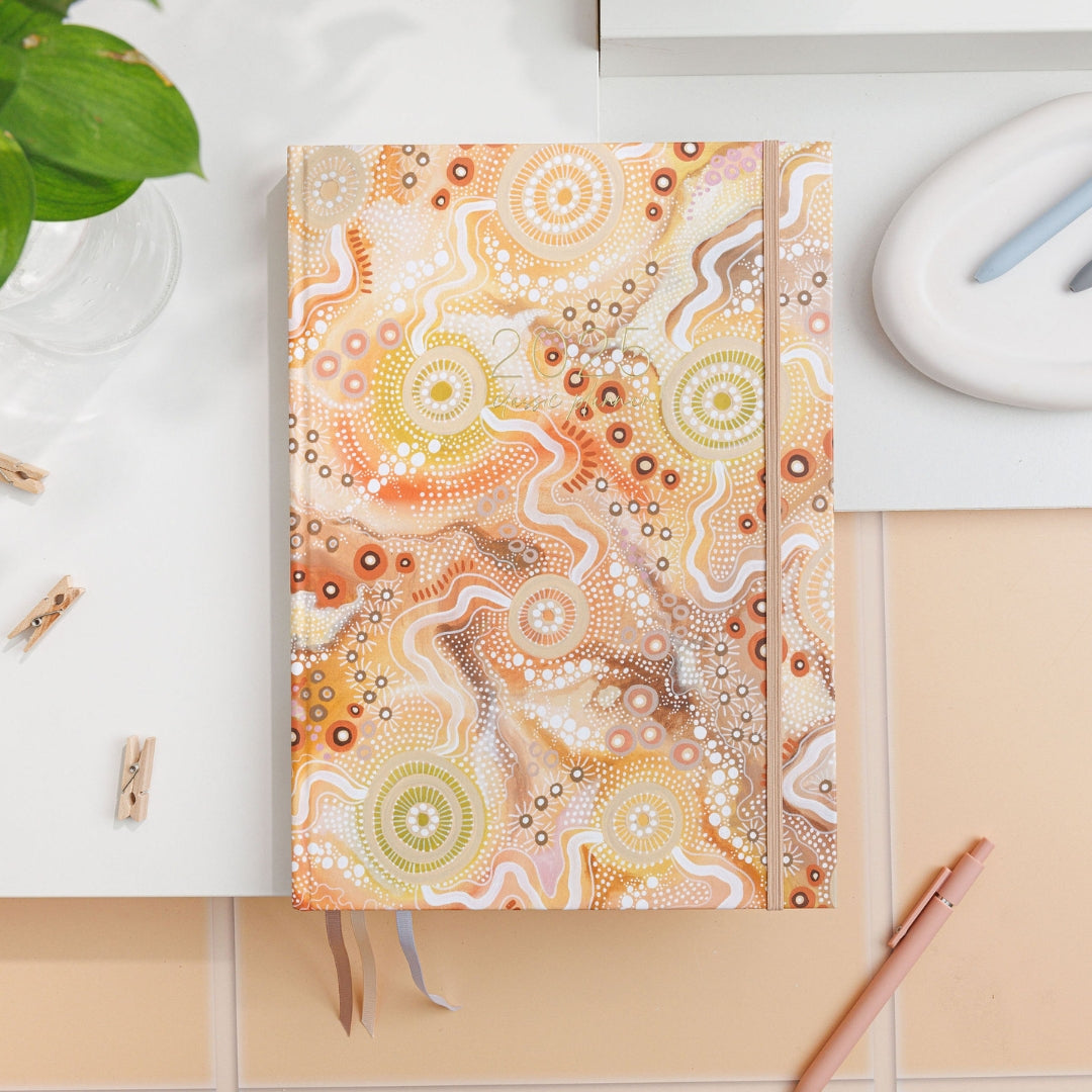 2025 Classic Weekly Teacher Planner named Women's Place, on a desk, featuring Aboriginal art in earthy oranges and browns.