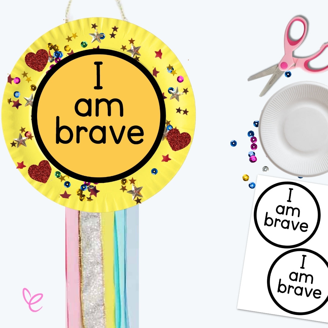  A yellow paper plate craft with "I am brave" written in bold letters, decorated with glittery stars, beads, and heart stickers. Colourful ribbons hang from the bottom, and scissors, a paper plate, and printable templates are visible in the background.