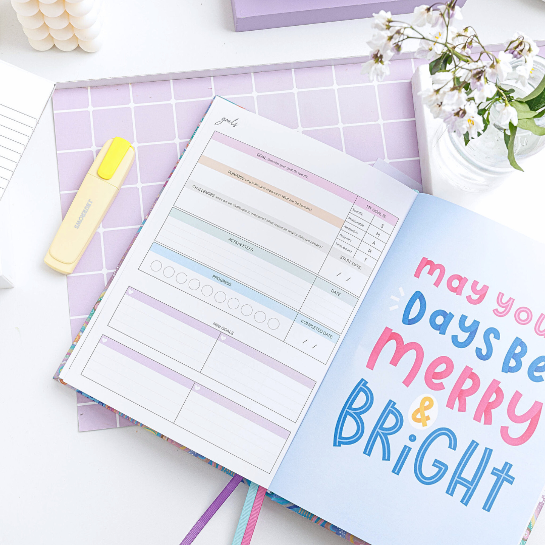 Open weekly planner showing a goals page on the left and a large-font quote on the right page reading 'May your days be merry & bright.'