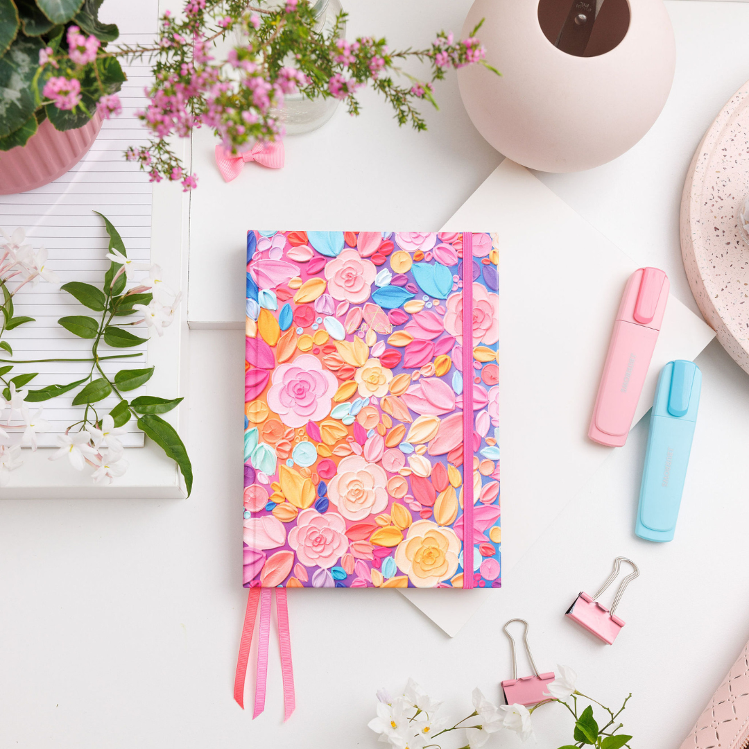 Awakening A5 Notepad with pink and red floral artwork, gold-foiled text, elastic enclosure, and ribbon bookmarks on a desk.