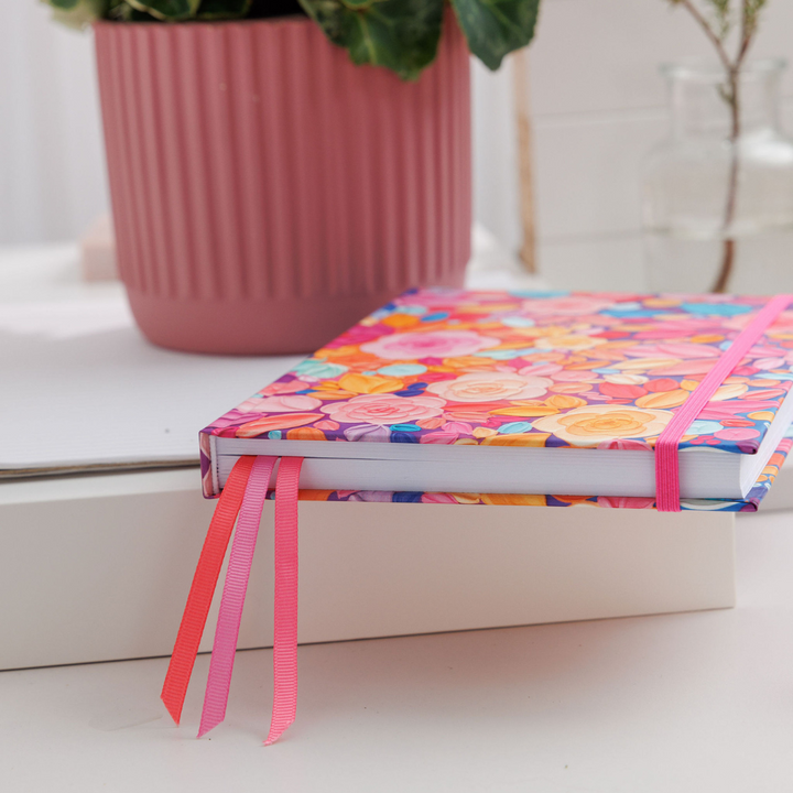 Awakening A5 Notepad lying on its side, showing the closed pages and ribbon bookmarks against the pink and red floral cover.