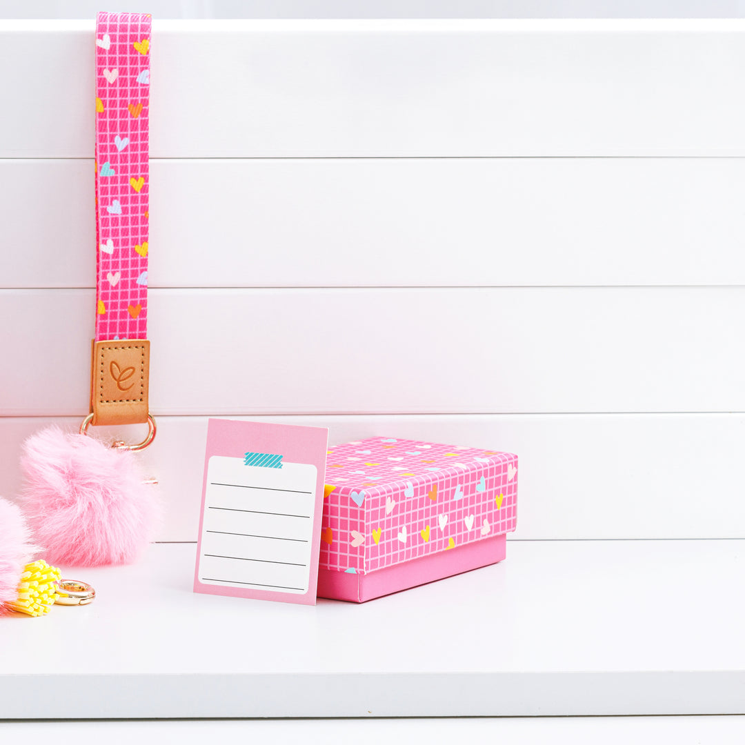 Bright pink wristlet with white grid and colourful heart pattern, featuring a light pink pom-pom, yellow tassel, and gold swivel clasp. A light brown faux leather patch displays the 'Mrs Edgar' logo. Comes with a matching gift box and thank-you card for teachers.