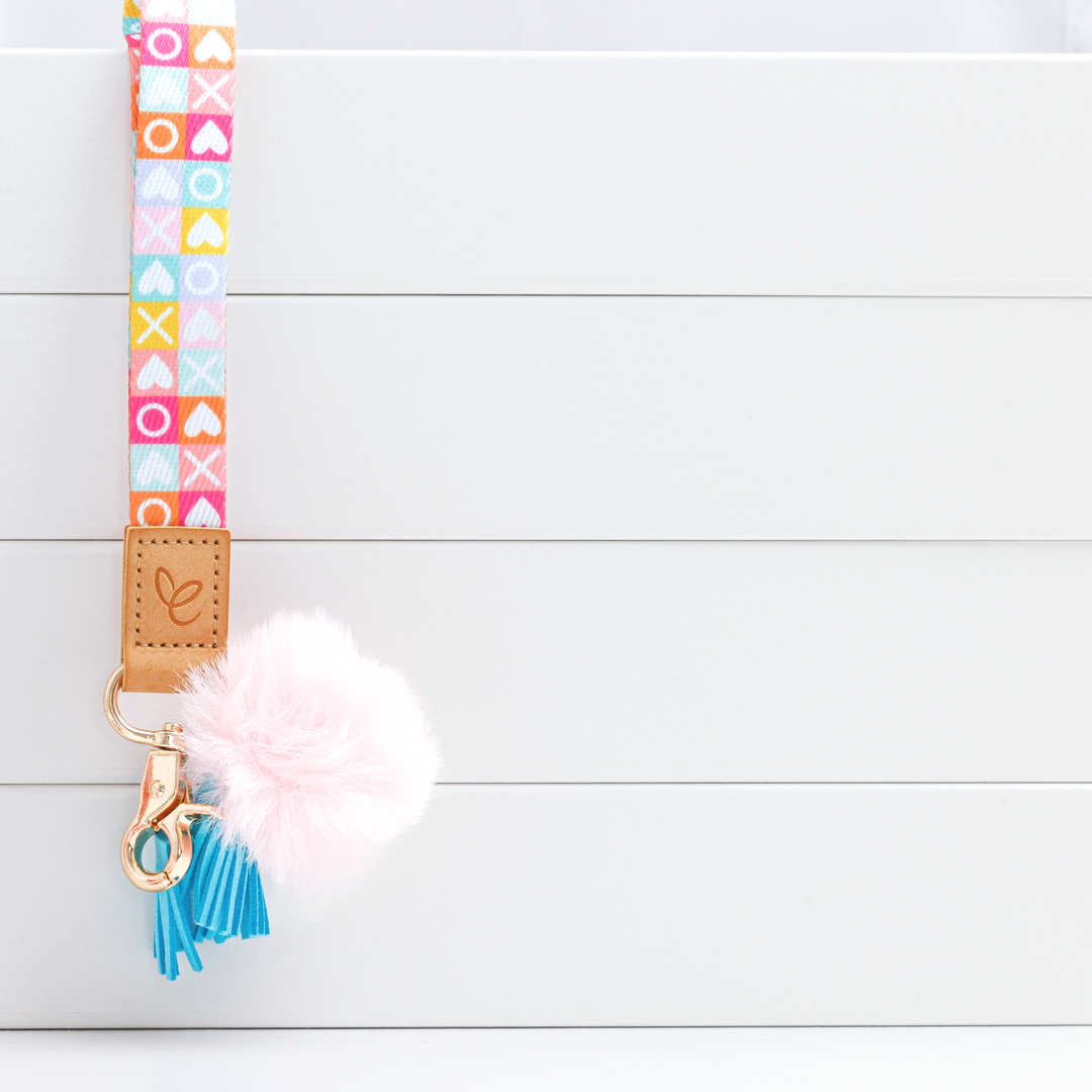 Colourful wristlet with alternating squares featuring white X, O, and hearts. Includes a light pink pom-pom, blue tassel, gold swivel clasp, and a light brown faux leather 'Mrs Edgar' logo patch.